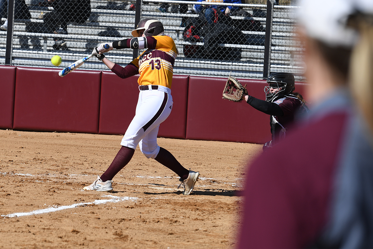 Coach Laura North watching the game as IHCC Student-Athlete swings bat towards incoming softball.